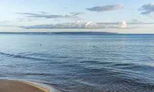 Sanfter Wellengang am Strand, im Hintergrund die Küstenlinie mit Häusern und Industrieanlagen, die an die Ostseeinseln erinnert. Ein einzelner Vogel schwimmt im ruhigen Wasser.