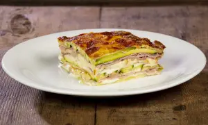 Una fetta di parmigiana di zucchine bianca alla napoletana, stratificata con prosciutto e formaggio fuso, su un piatto bianco.
