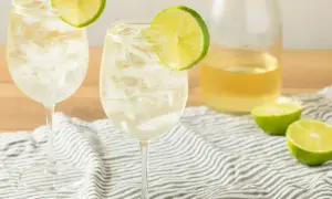 Orzeźwiający szprycer drink z lodem i plasterkiem limonki, obok butelka białego wina i połówki limonki.