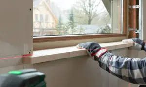 Precyzyjny montaż parapetu drewnianego z użyciem poziomicy laserowej. Ręce w rękawicach przytrzymują deskę, a czerwone linie lasera wyznaczają idealny poziom.