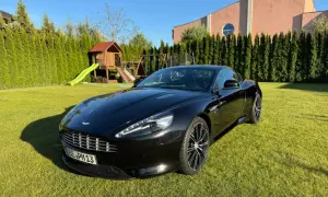 Czarny Aston Martin DBS lśni na trawniku przed domem. W tle plac zabaw i wysokie tuje.