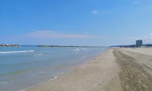 Spiaggia del lido di Fermo con mare calmo, frangiflutti in pietra e un palazzo sullo sfondo.