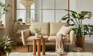Przytulny salon w stylu cottage: beżowa kanapa, drewniany stolik, dużo roślin i pleciony klosz lampy tworzą ciepłą atmosferę.