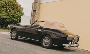 Svart, klassisk brittisk sportbil med beige sufflett parkerad vid en gul vägg, bredvid ett apelsinträd.