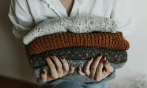 Dłonie trzymają stos ciepłych swetrów. Widać różne odcienie szarości i brązu, a każdy sweter to miękka tkanina wełniana.