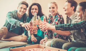 Grupa przyjaciół wznosi toast butelkami piwa, śmiejąc się i jedząc pizzę. Nawet trudne pytania nie psują im nastroju.