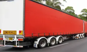 Un long camion 60 tonnes, avec une remorque rouge vif, roule sur une route bordée d'arbres.