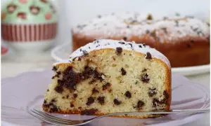 Una fetta di torta soffice con tante gocce di cioccolato, cosparsa di zucchero a velo, su un piatto lilla con una forchetta.