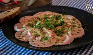 Saure Wurst mit Zwiebeln, Radieschen und Schnittlauch auf schwarzem Teller, dazu Brot.