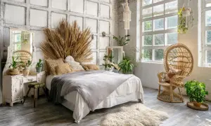 Sypialnia w stylu boho z łóżkiem, wiklinowym fotelem i dużą ilością roślin. Dominują naturalne materiały i ciepłe, ziemiste odcienie, tworząc przytulny klimat.