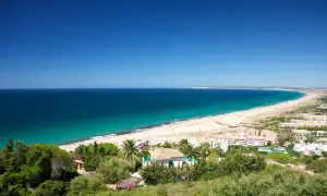 Vista panorámica del pueblo y paseo marítimo de Zahara de los Atunes, con su extensa playa de arena dorada y el mar azul turquesa.