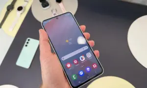 Ręka trzyma smartfon Samsung, na ekranie widoczna pogoda i ikony aplikacji. W tle leżą zegarki i słuchawki. Czas przed ekranem Samsung upływa przyjemnie.