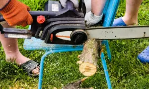Osoba w pomarańczowej rękawicy używa piły łańcuchowej do cięcia drewna na prowizorycznym stojaku. To prosty sposób, jak zrobić stojak do cięcia drewna.