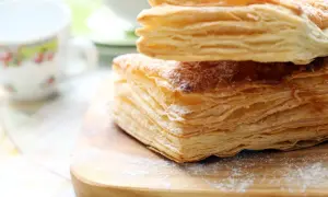 Stos puszystego ciasta francuskiego, idealnego do nauki, jak zrobić ciasto francuskie. Posypane cukrem pudrem, na drewnianej desce.