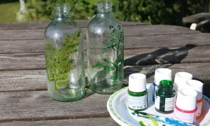 Två glasflaskor målas med gröna växtmotiv. Bredvid står färgtuber och penslar, redo att måla ljuslykta.