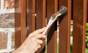 Dłoń maluje pędzlem brązowy płot drewniany. Idealny wybór, czym pomalować płot drewniany, by był piękny i chroniony.