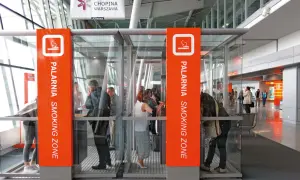 Szklane kabiny palarni na lotnisku z pomarańczowymi oznaczeniami i ludźmi w środku.