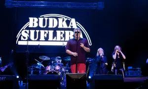 Budka Suflera na scenie. Wokalista w okularach śpiewa, a obok niego dwie wokalistki. W tle perkusja i logo zespołu.