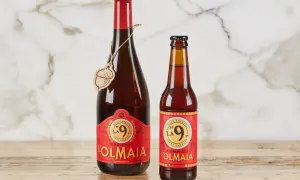 Due bottiglie di birra ambrata "La 9" del Birrificio L'Olmaia, una da 750ml e una più piccola, su un tavolo di legno.