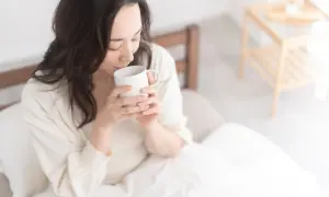 Donna con capelli scuri beve acqua calda al mattino, seduta a letto, avvolta in una coperta bianca.