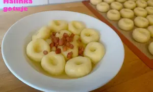 Pyszne kluski śląskie z okrasą z boczku, gotowe do podania. Na blacie widać kolejne, przygotowane do gotowania.
