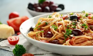 Un piatto di spaghetti alla puttanesca, conditi con pomodoro, olive e capperi, guarnito con prezzemolo e parmigiano.