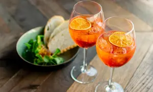 Zwei Gläser mit einem orangen Aperitif, garniert mit Orangenscheiben, daneben ein Schälchen mit Salat und Brot.