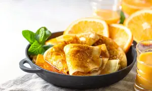Złociste crepes suzette w sosie pomarańczowym, udekorowane miętą i plasterkami pomarańczy, obok szklanki soku.