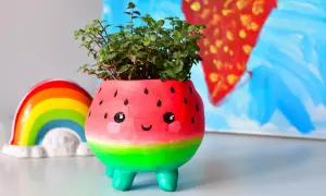 Urocza doniczka diy w kształcie arbuza z uśmiechniętą buźką, pełna zielonych roślinek. Obok tęczowa ozdoba.
