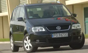 Czarny VW Touran z przodu, widoczne reflektory i żarówki. Kierowca za kierownicą.
