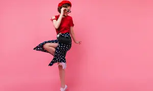 Urocza dziewczyna w stylu pin-up girl w czerwonej bluzce, spódnicy w grochy i czerwonym beretcie, skacze z radości na różowym tle.