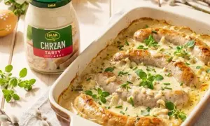 Biała kiełbasa w sosie chrzanowym, zapiekana w naczyniu, z dodatkiem ziół. Obok słoik sosu chrzanowego.