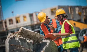Operai con caschi gialli e giubbotti ad alta visibilità discutono la pianificazione progetto su un cantiere, esaminando documenti e smartphone.