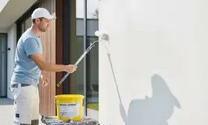 Mann streicht mit einer Fassadenrolle eine weiße Wand. Ein Eimer mit Farbe steht daneben.