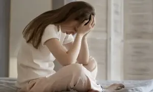 Kobieta siedzi na łóżku, obejmując głowę rękami. Jej postawa sugeruje walkę z **zespołem stresu pourazowego**.