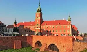 Zamek Królewski w Warszawie, z czerwoną fasadą i zieloną wieżą, widziany zza ceglanego mostu.