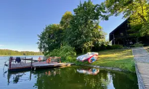 Drewniane pomosty nad spokojnym jeziorem raczyńskim, obok leżaków i dwóch łódek. W tle drewniany dom i bujna zieleń.