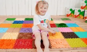 Uśmiechnięta dziewczynka bawi się na kolorowej macie sensorycznej dla niemowląt, odkrywając różne faktury i kształty.