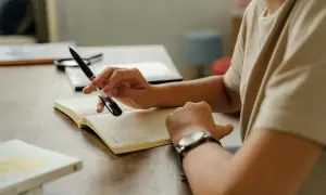 Osoba z długopisem w dłoni i zegarkiem na ręku zastanawia się, jak napisać pismo do prokuratury, notując w dzienniku.