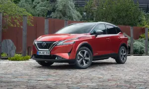 Czerwony Nissan Qashqai 2025 z czarnym dachem, zaparkowany na brukowanej nawierzchni, otoczony zielenią i industrialnym ogrodzeniem.