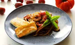 Makaron z halloumi na talerzu, z sosem pomidorowym, oliwkami i bazylią. Obok leżą świeże pomidory.