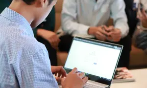Mężczyzna w niebieskiej koszuli pracuje na laptopie. W tle widać innych ludzi, być może to spotkanie z kontrahentem.