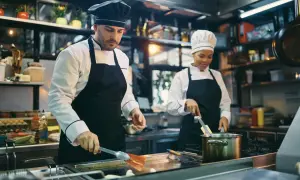 Dwóch kucharzy w kuchni restauracyjnej przygotowuje posiłek. Mężczyzna z przodu używa łopatki, kobieta z tyłu miesza coś w garnku. Kod zawodu kucharz.
