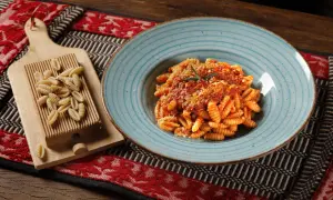 Gnocchetti sardi conditi con sugo rosso e parmigiano, accanto a una spianatoia con pasta fresca.