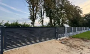 Nowoczesne, ciemnoszare ogrodzenie z przesuwną bramą. Widać kamienny murek i drzewa w tle. Odległość ogrodzenia od drogi gminnej wewnętrznej jest niewielka.