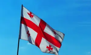 Flaga Gruzji powiewa na tle błękitnego nieba. Biały materiał ozdobiony jest dużym czerwonym krzyżem i czterema mniejszymi krzyżami w rogach.