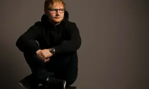 Ed Sheeran w czarnej bluzie z kapturem i okularach siedzi ze skrzyżowanymi nogami.