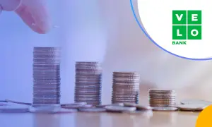 Ręka dodaje monetę do rosnącej stosu, symbolizując wzrost kapitału. VeloBank oprocentowanie lokat rośnie wraz z Twoimi oszczędnościami.