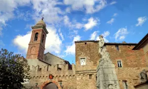 Poggio San Marcello: campanile in mattoni, un arco fortificato e una statua di bronzo sotto un cielo azzurro con nuvole bianche.