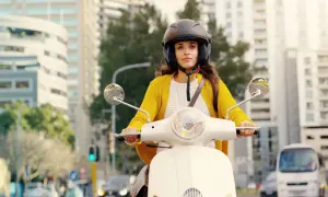 Kobieta w kasku jedzie białym motorowerem przez miasto. Czy na motorower trzeba mieć prawo jazdy? To pytanie nurtuje wielu.